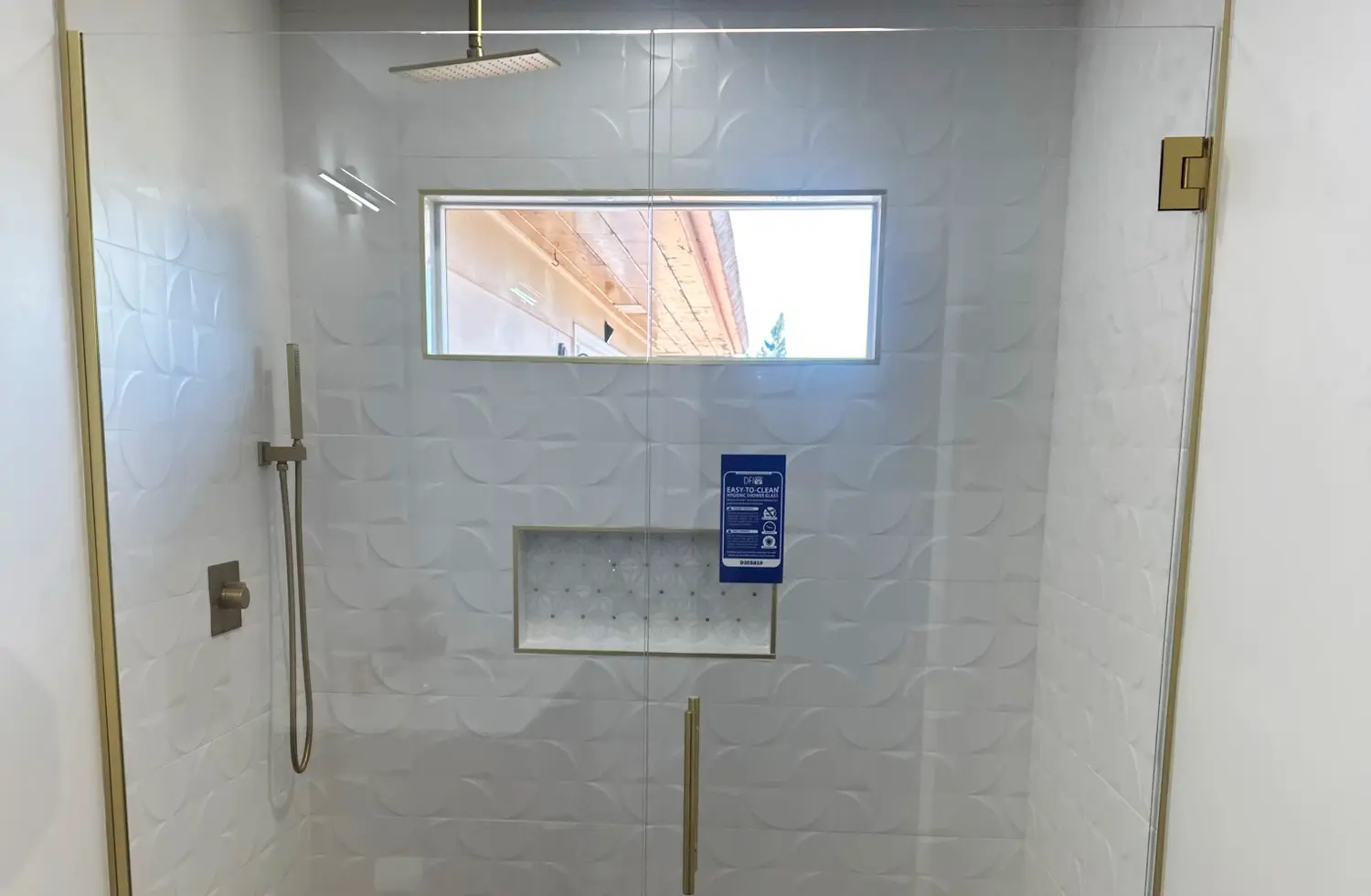 Newly installed frameless glass shower featuring white patterned tile walls, gold shower fixtures, and a recessed shelf